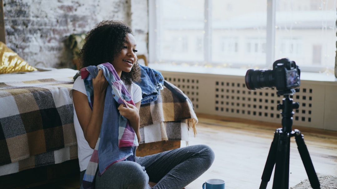Attractive african girl sitting near bed recording video blog about her wardrobe for travel with dslr camera at home
