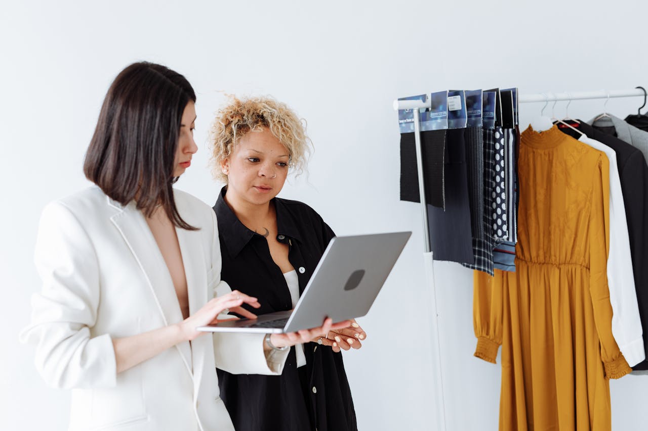 Two women fashion designers collaborating with a laptop in a modern workspace with clothing samples.